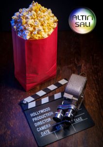 Popcornia punaisessa pussissa sekä elokuvaleikasin ja vintage-elokuvakamera pöydällä, Altin salin logo taustalla.